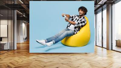 Full body young Indian man wears shirt white t-shirt casual clothes sit in bag chair hold in hand play pc game with joystick console isolated on plain pastel blue cyan background. Lifestyle concept. Wall mural