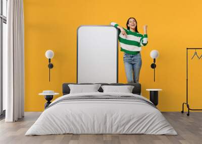 Full body young latin woman wear casual cozy green knitted sweater big huge blank screen mobile cell phone smartphone with area do winner gesture isolated on plain yellow background studio portrait. Wall mural