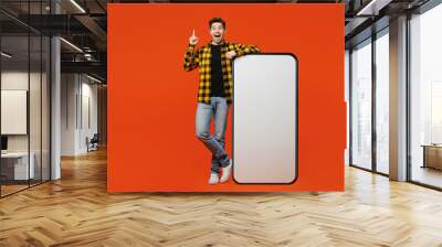 Full body young man he wear yellow checkered shirt black t-shirt big huge blank screen mobile cell phone smartphone with workspace area point index finger up isolated on plain red orange background. Wall mural