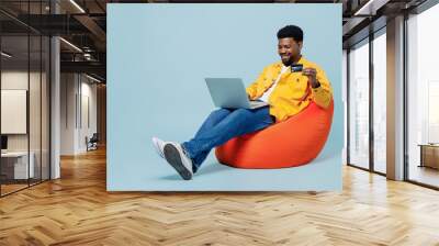 Full body young man of African American ethnicity in yellow shirt sit in bag chair use laptop computer credit bank card shopping online order delivery isolated on plain pastel light blue background Wall mural