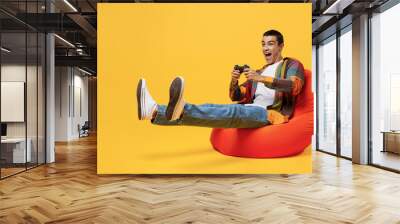 Full body young middle eastern man 20s he wear casual shirt white t-shirt sit in bag chair hold in hand play pc game with joystick console isolated on plain yellow background People lifestyle concept. Wall mural