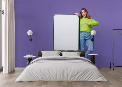 Full body young smiling fun woman 30s she wear casual green knitted sweater stand point finger on big huge blank screen mobile cell phone with area isolated on plain pastel purple background studio. Wall mural