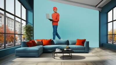 Full body young smiling happy african american man 20s in orange shirt hat use hold laptop pc computer look back aside isolated on plain pastel light blue background studio. People lifestyle concept Wall mural