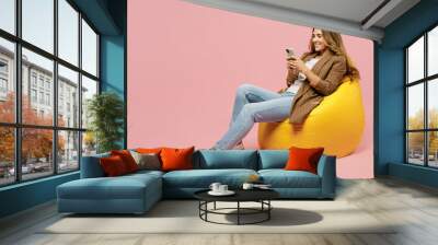 Full body young successful employee business woman 30s she wear casual brown classic jacket sit in bag chair hold in hand use mobile cell phone isolated on plain pastel light pink background studio. Wall mural