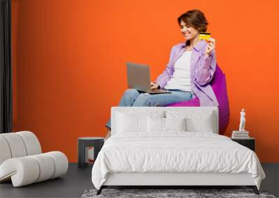 Full body young woman wear purple shirt white t-shirt casual clothes sit in bag chair using laptop pc computer hold credit bank card shopping online booking tour isolated on plain orange background. Wall mural