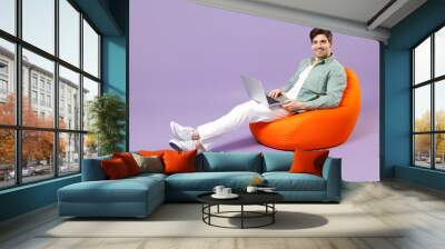 Full length fun smiling man 20s wear casual mint shirt white t-shirt sitting in orange bean bag chair use laptop pc computer chat isolated on purple color background studio People lifestyle concept Wall mural