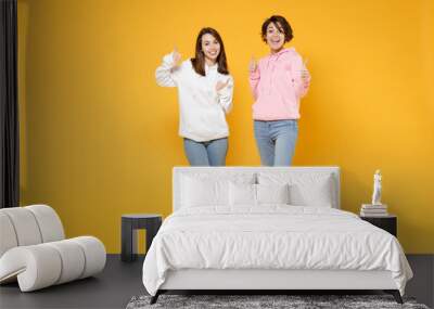 Full length of excited cheerful two young women friends 20s wearing basic white pink hoodies standing showing thumbs up like gesture looking camera isolated on yellow color background studio portrait. Wall mural