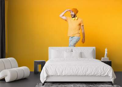 Full length of excited surprised young man wearing basic casual t-shirt headphones hat holding hand at forehead looking far away distance isolated on bright yellow colour background, studio portrait. Wall mural