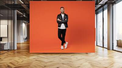 Full length of smiling attractive young bearded man 20s wearing basic white t-shirt black leather jacket standing holding hands crossed looking camera isolated on orange background studio portrait. Wall mural