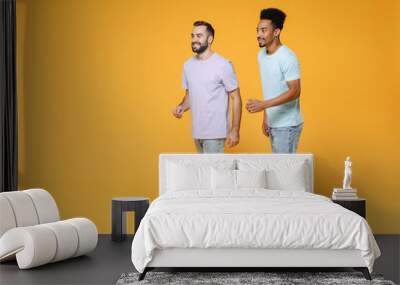 Full length of smiling young two friends european african american men 20s wearing purple blue casual t-shirts walking going looking aside isolated on bright yellow colour background studio portrait. Wall mural