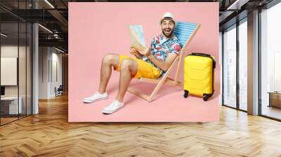 Full length of surprised young traveler tourist man in summer clothes hat sit on deck chair hold city map isolated on pink background studio. Passenger travel on weekend. Air flight journey concept. Wall mural