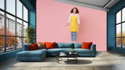 Full length portrait of smiling young brunette woman housewife 20s wearing yellow apron jumping having fun while doing housework isolated on pastel pink colour background studio. Housekeeping concept. Wall mural