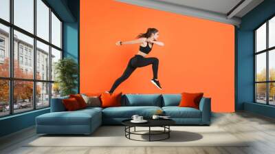 Full length side view of portrait of young fitness sporty woman 20s wearing black sportswear posing training working out jumping like running looking aside isolated on orange color background studio. Wall mural