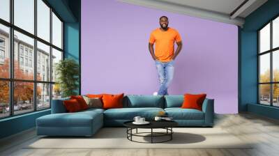 Full length side view of smiling young african american man 20s wearing basic casual orange t-shirt holding hands in pockets looking camera isolated on pastel violet colour background studio portrait. Wall mural