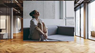 Full length side view young satisfied dreamful relaxed caucasian woman 20s wearing casual white clothes sit on soft grey sofa indoors apartment hugging knees. Resting on weekends staying home concept Wall mural