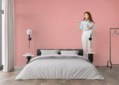 Full length side view young surprised redhead woman in blue shirt pants using mobile cell phone point index finger aside on workspace area mock up isolated on pastel pink background studio portrait Wall mural