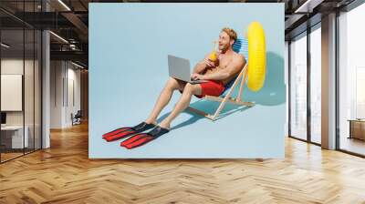 Full length young man 20s in red shorts swimsuit sit in deckchair inflatable ring hold cocktail use laprop pc computer work isolated on pastel blue background Summer vacation sea rest sun tan concept Wall mural