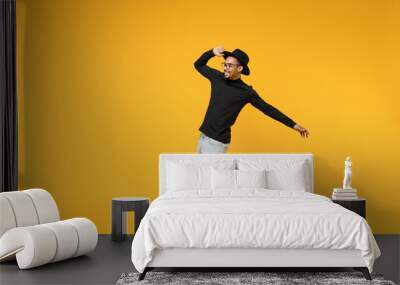 Full length young smiling fashionable fun african man 20s wearing stylish black hat shirt eyeglasses stand on toes dancing leaning back dancing isolated on yellow orange background studio portrait Wall mural