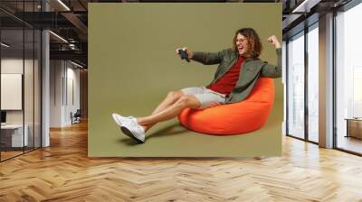 Full size body length fun young brunet curly man 20s wear khaki shirt sit in bag chair play pc game with joystick console doing winner gesture isolated on plain olive green background studio portrait Wall mural