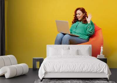 Full size body length young ginger chubby woman 20s wears green shirt sit in bag chair hold use work on laptop pc computer get video call wave hand isolated on plain yellow background studio portrait. Wall mural