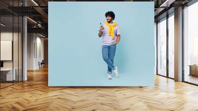 Full size young bearded Indian man 20s years old wear white t-shirt hold in hand use mobile cell phone typing browsing chatting send sms isolated on plain pastel light blue background studio portrait Wall mural