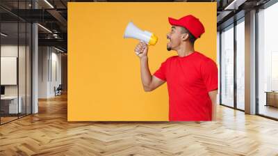 Fun crazy delivery employee african man in red cap blank t-shirt uniform workwear work courier service concept hold scream in megaphone announces sale Hurry up isolated on yellow background studio Wall mural