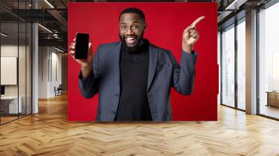 Funny young african american business man 20s in classic jacket suit standing hold mobile phone with blank empty screen pointing index finger aside up isolated on red color background studio portrait. Wall mural