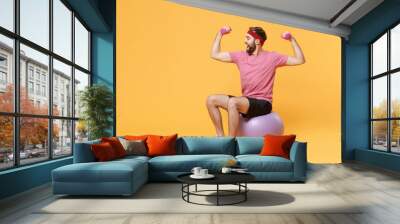 Funny young bearded fitness sporty guy sportsman in headband t-shirt in home gym isolated on yellow background. Workout sport motivation lifestyle concept. Sit on fitball doing exercise with dumbbell. Wall mural