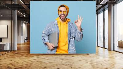 Funny young hipster guy in fashion jeans denim clothes posing isolated on pastel blue background in studio. People lifestyle concept. Mock up copy space. Hold laptop pc computer, showing OK gesture. Wall mural