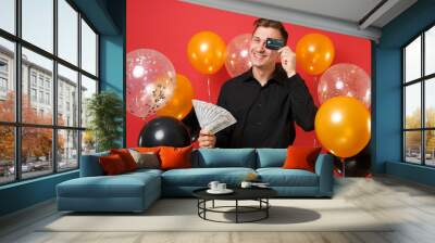 Funny young man in black classic shirt hold bundle lots of dollars cash money covering eye with credit card on bright red background air balloons. Happy New Year birthday mockup holiday party concept. Wall mural