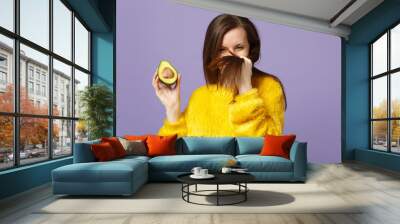 Funny young woman in fur sweater touching hair holding in hand half of fresh ripe green avocado isolated on violet pastel background. People vivid lifestyle relax vacation concept. Mock up copy space. Wall mural