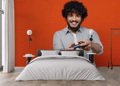 Gambling overjoyed cheerful excited jubilant young bearded Indian man 20s years old wear blue shirt hold in hand play pc game with joystick console isolated on plain orange background studio portrait Wall mural