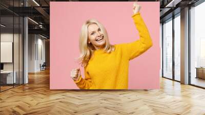 Happy cheerful elderly gray-haired blonde woman lady 40s 50s years old in yellow casual sweater standing doing winner gesture clenching fists isolated on pastel pink color background studio portrait. Wall mural