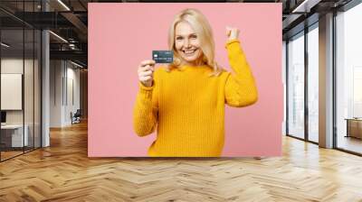 Happy elderly gray-haired blonde woman lady 40s 50s years old wearing yellow basic sweater standing hold credit bank card doing winner gesture isolated on pastel pink color background studio portrait. Wall mural