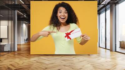 Happy excited cheerful beautiful african american young brunette woman 20s wears green shirt hold point on gift certificate coupon voucher card for storeisolated on yellow background studio portrait Wall mural