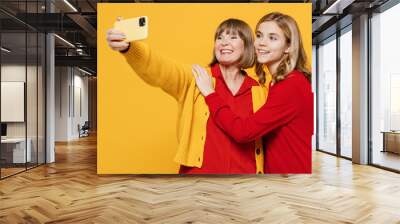 Happy fun woman 50s in red shirt with teenager girl 12-13 years old Grandmother granddaughter do selfie shot on mobile cell phone post photo on social network hug isolated on plain yellow background Wall mural