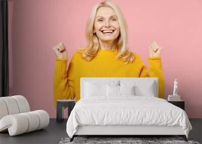 Happy joyful elderly gray-haired blonde woman lady 40s 50s years old in yellow casual sweater standing doing winner gesture clenching fists isolated on pastel pink color background studio portrait. Wall mural