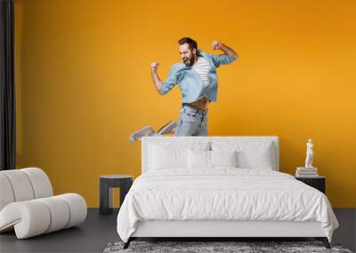 Happy young bearded man in casual blue shirt posing isolated on yellow orange background, studio portrait. People sincere emotions lifestyle concept. Mock up copy space. Jumping doing winner gesture. Wall mural