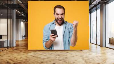 Happy young man in casual blue shirt posing isolated on yellow orange wall background studio portrait. People lifestyle concept. Mock up copy space. Hold mobile phone, doing winner gesture, screaming. Wall mural