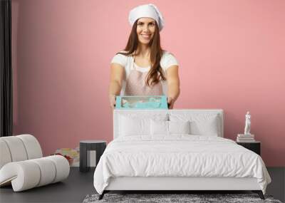 Housewife female chef cook confectioner or baker in apron white t-shirt, toque chefs hat giving cake cupcake in box table isolated on pink pastel background in studio. Mock up copy space food concept. Wall mural