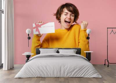 Joyful young brunette woman girl in yellow sweater posing isolated on pastel pink background studio portrait. People lifestyle concept. Mock up copy space. Hold gift certificate, doing winner gesture. Wall mural