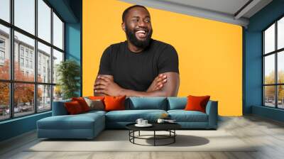 Laughing african american man guy football fan in casual black t-shirt isolated on yellow background studio portrait. People lifestyle concept. Mock up copy space. Holding hands crossed looking aside. Wall mural