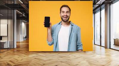 Laughing young man in casual blue shirt posing isolated on yellow orange wall background, studio portrait. People lifestyle concept. Mock up copy space. Holding mobile phone with blank empty screen. Wall mural