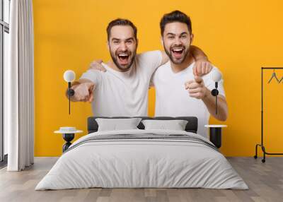 Laughing young men guys friends in white blank empty t-shirts posing isolated on yellow orange background studio portrait. People lifestyle concept. Mock up copy space. Point index fingers on camera. Wall mural