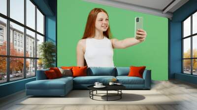 Little redhead kid girl 12-13 years old wearing white tank shirt doing selfie shot on mobile cell phone post photo on social network isolated on plain green background. Childhood lifestyle concept Wall mural