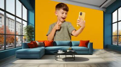 Little small smiling boy 6-7 years old in green t-shirt doing selfie shot on mobile cell phone point finger on camera isolated on plain yellow background. Mother's Day love family lifestyle concept. Wall mural