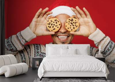 Merry cheerful fun joyful young man wear warm cozy Christmas sweater Santa hat posing cover eyes with cookies biscuits isolated on plain red background Happy New Year 2023 celebration holiday concept Wall mural