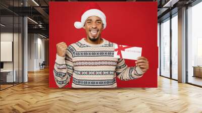 Merry fun young man wear Christmas sweater Santa hat posing hold gift certificate coupon voucher card for store do winner gesture isolated on plain red background. Happy New Year 2023 holiday concept. Wall mural