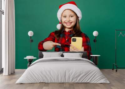 Merry happy fun little child kid girl 6-7 years old wear red dress Christmas hat posing hold use point finger on mobile cell phone isolated on plain dark green background. Happy New Year 2023 concept. Wall mural