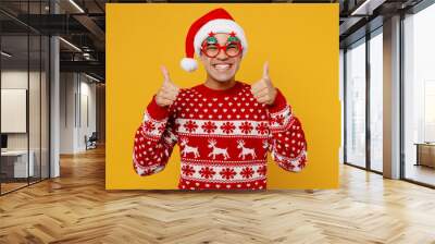 Merry satisfied fun young man wear knitted christmas sweater Santa hat glasses posing show thumb up like gesture isolated on plain yellow background. Happy New Year 2023 celebration holiday concept. Wall mural